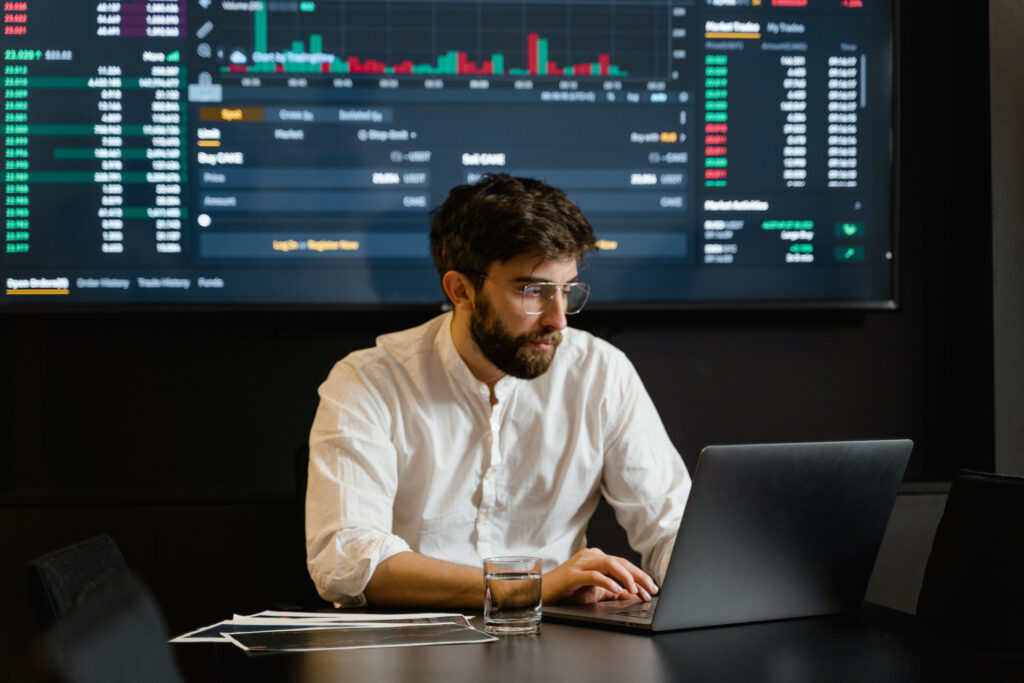 Eine Person sitzt am Laptop und arbeitet. Hinter ihm ein großer Bildschirm mit Analysedaten.