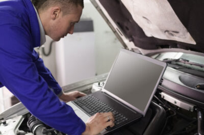 Ein Mechaniker arbeitet bei geöffneter Motorhaube am Auto mit einem Laptop.