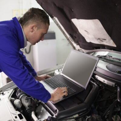 Ein Mechaniker arbeitet bei geöffneter Motorhaube am Auto mit einem Laptop.