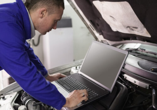 Ein Mechaniker arbeitet bei geöffneter Motorhaube am Auto mit einem Laptop.