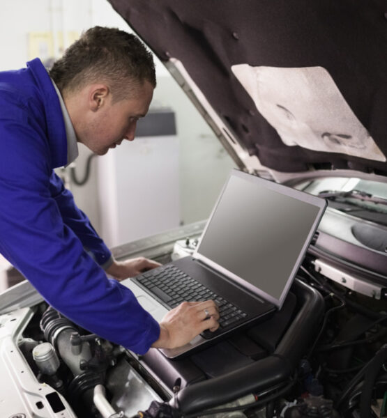 Ein Mechaniker arbeitet bei geöffneter Motorhaube am Auto mit einem Laptop.