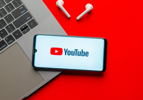 Ein Smartphone mit dem YouTube-Logo auf dem Display liegt neben einem Laptop und kabellosen Ohrhörern auf einem roten Hintergrund.
