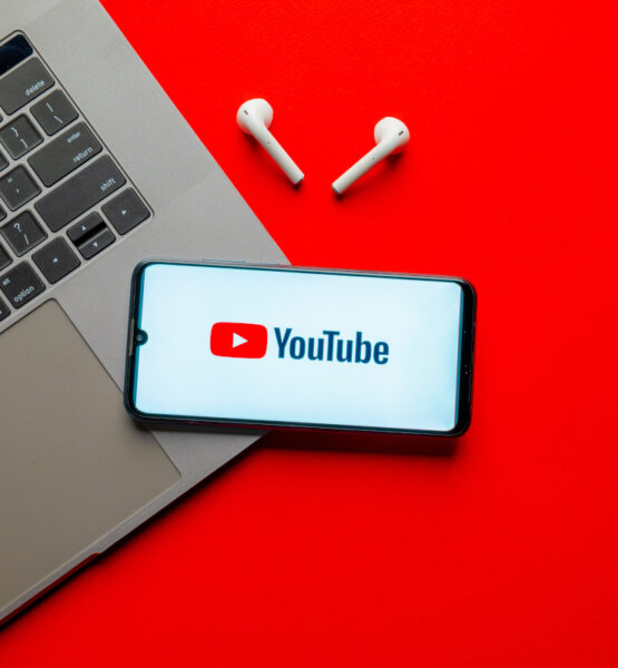 Ein Smartphone mit dem YouTube-Logo auf dem Display liegt neben einem Laptop und kabellosen Ohrhörern auf einem roten Hintergrund.