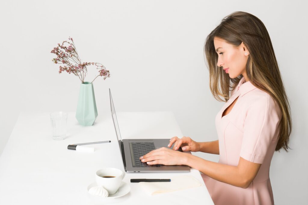 Eine junge Frau arbeitet an ihrem Laptop. Auf dem weissen Tisch steht eine kleine Blumenvase und eine Tasse Kaffee.