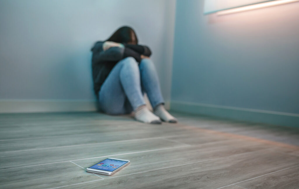 Eine Person sitzt zusammengekauert in der Ecke ihres Zimmers. Etwas von ihr entfernt liegt ein Smartphone auf dem Boden.
