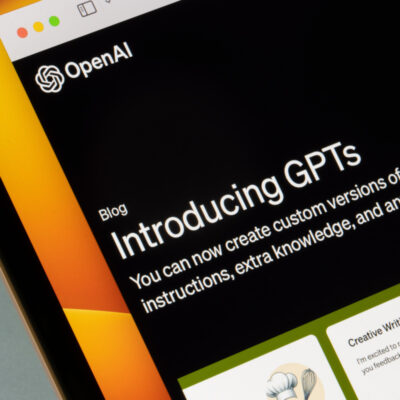 Ein Computerbildschirm zeigt eine Webseite von OpenAI mit einem Blogbeitrag mit dem Titel "Introducing GPTs". Der Text darunter erklärt, dass man nun benutzerdefinierte Versionen von ChatGPT erstellen kann, die Anweisungen, zusätzliches Wissen und eine beliebige Kombination von Fähigkeiten kombinieren. Im unteren Bereich des Bildschirms sind weitere Inhalte zu sehen, darunter ein Abschnitt mit der Überschrift "Creative Writing Coach".