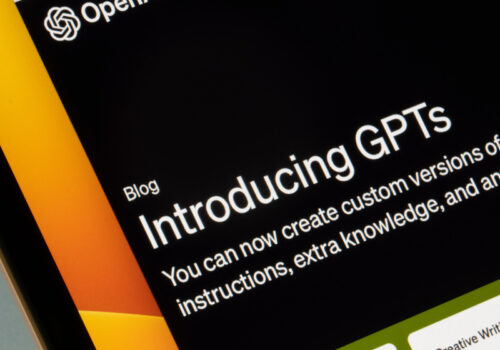 Ein Computerbildschirm zeigt eine Webseite von OpenAI mit einem Blogbeitrag mit dem Titel "Introducing GPTs". Der Text darunter erklärt, dass man nun benutzerdefinierte Versionen von ChatGPT erstellen kann, die Anweisungen, zusätzliches Wissen und eine beliebige Kombination von Fähigkeiten kombinieren. Im unteren Bereich des Bildschirms sind weitere Inhalte zu sehen, darunter ein Abschnitt mit der Überschrift "Creative Writing Coach".