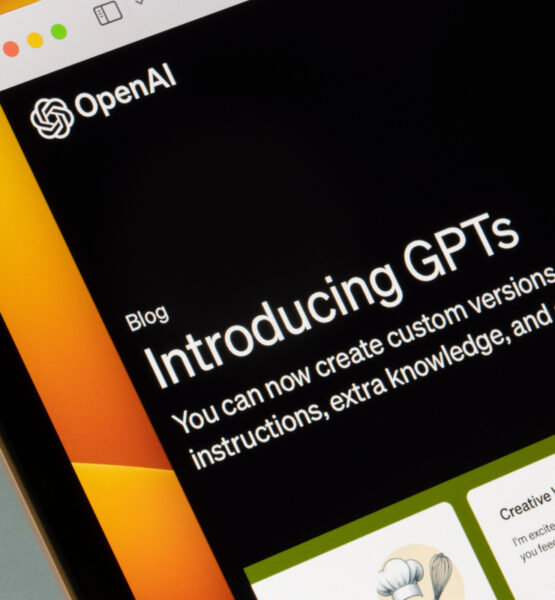 Ein Computerbildschirm zeigt eine Webseite von OpenAI mit einem Blogbeitrag mit dem Titel "Introducing GPTs". Der Text darunter erklärt, dass man nun benutzerdefinierte Versionen von ChatGPT erstellen kann, die Anweisungen, zusätzliches Wissen und eine beliebige Kombination von Fähigkeiten kombinieren. Im unteren Bereich des Bildschirms sind weitere Inhalte zu sehen, darunter ein Abschnitt mit der Überschrift "Creative Writing Coach".