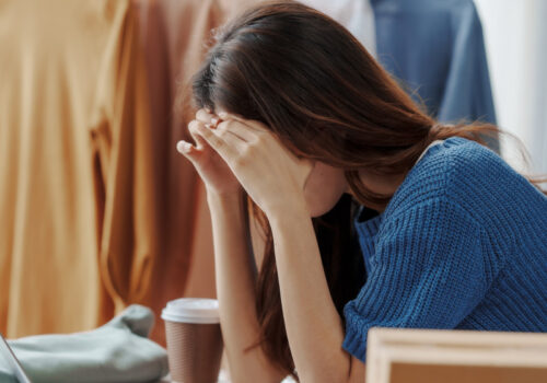 Eine Frau stützt den Kopf vor dem Laptop erschöpft in ihre Hände, offenbar mit Kopfschmerzen. Im Hintergrund ist eine Kleiderstanke mit mehreren Oberteilen zu sehen.