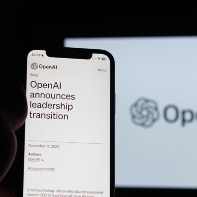 Ein Smartphone, das in der Hand gehalten wird, zeigt einen Blogbeitrag von OpenAI mit der Überschrift "OpenAI announces leadership transition" auf dem Bildschirm. Impliziert werden Veränderungen in der KI-Entwicklung. Im Hintergrund ist unscharf das Logo von OpenAI auf einem größeren Bildschirm zu sehen. Es ist dunkel um das Telefon herum, was den Bildschirminhalt hervorhebt.