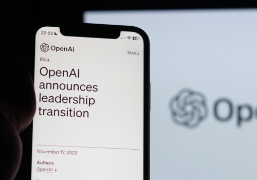 Ein Smartphone, das in der Hand gehalten wird, zeigt einen Blogbeitrag von OpenAI mit der Überschrift "OpenAI announces leadership transition" auf dem Bildschirm. Impliziert werden Veränderungen in der KI-Entwicklung. Im Hintergrund ist unscharf das Logo von OpenAI auf einem größeren Bildschirm zu sehen. Es ist dunkel um das Telefon herum, was den Bildschirminhalt hervorhebt.