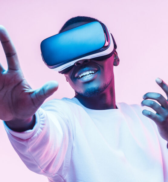 Ein lächelnder Mann trägt eine Virtual-Reality-Brille und streckt seine Hände aus, als würde er etwas in der virtuellen Realität berühren oder darauf reagieren. Er scheint begeistert und fasziniert von der Erfahrung zu sein. Der Hintergrund ist in weiches rosa und blaues Licht getaucht, was eine futuristische Atmosphäre schafft.