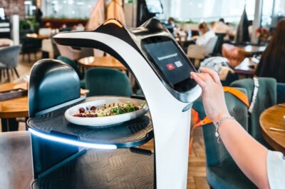 Eine Frau bedient ein modernes, digitales Selbstbedienungs-Terminal in einem Restaurant, auf dem möglicherweise die Bestellung aufgegeben oder bezahlt wird. Vor ihr auf dem Tisch steht ein Teller mit Salat. Das Terminal hat ein futuristisches Design mit blauen Leuchtelementen. Im Hintergrund ist das Innere des Restaurants mit weiteren Gästen und Tischen zu erkennen Es handelt sich um einen Küchenroboter.