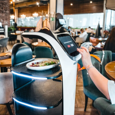 Eine Frau bedient ein modernes, digitales Selbstbedienungs-Terminal in einem Restaurant, auf dem möglicherweise die Bestellung aufgegeben oder bezahlt wird. Vor ihr auf dem Tisch steht ein Teller mit Salat. Das Terminal hat ein futuristisches Design mit blauen Leuchtelementen. Im Hintergrund ist das Innere des Restaurants mit weiteren Gästen und Tischen zu erkennen Es handelt sich um einen Küchenroboter.