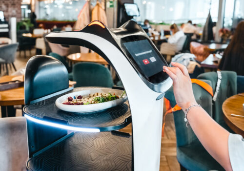 Eine Frau bedient ein modernes, digitales Selbstbedienungs-Terminal in einem Restaurant, auf dem möglicherweise die Bestellung aufgegeben oder bezahlt wird. Vor ihr auf dem Tisch steht ein Teller mit Salat. Das Terminal hat ein futuristisches Design mit blauen Leuchtelementen. Im Hintergrund ist das Innere des Restaurants mit weiteren Gästen und Tischen zu erkennen Es handelt sich um einen Küchenroboter.