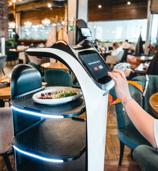 Eine Frau bedient ein modernes, digitales Selbstbedienungs-Terminal in einem Restaurant, auf dem möglicherweise die Bestellung aufgegeben oder bezahlt wird. Vor ihr auf dem Tisch steht ein Teller mit Salat. Das Terminal hat ein futuristisches Design mit blauen Leuchtelementen. Im Hintergrund ist das Innere des Restaurants mit weiteren Gästen und Tischen zu erkennen Es handelt sich um einen Küchenroboter.