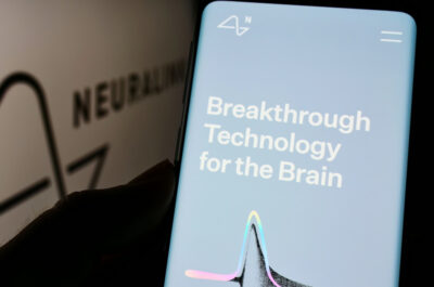 Ein Smartphone wird in der Hand gehalten und zeigt eine Anzeige mit dem Text "Breakthrough Technology for the Brain". Im Hintergrund ist das Logo von Neuralink, einem Unternehmen, das sich auf die Entwicklung von Neurotechnologie spezialisiert hat, teilweise sichtbar. Das Logo erscheint unscharf und hinter dem Smartphone, was darauf hindeutet, dass der Fokus auf dem Bildschirm des Smartphones liegt. Der Bildschirm ist hellblau mit weißem Text und einem grafischen Element in der Mitte, das eine stilisierte Darstellung einer Neuronen- oder Synapsenstruktur sein könnte, mit Farbakzenten in Rosa und Grün an den Enden.