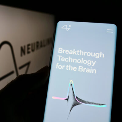 Ein Smartphone wird in der Hand gehalten und zeigt eine Anzeige mit dem Text "Breakthrough Technology for the Brain". Im Hintergrund ist das Logo von Neuralink, einem Unternehmen, das sich auf die Entwicklung von Neurotechnologie spezialisiert hat, teilweise sichtbar. Das Logo erscheint unscharf und hinter dem Smartphone, was darauf hindeutet, dass der Fokus auf dem Bildschirm des Smartphones liegt. Der Bildschirm ist hellblau mit weißem Text und einem grafischen Element in der Mitte, das eine stilisierte Darstellung einer Neuronen- oder Synapsenstruktur sein könnte, mit Farbakzenten in Rosa und Grün an den Enden.
