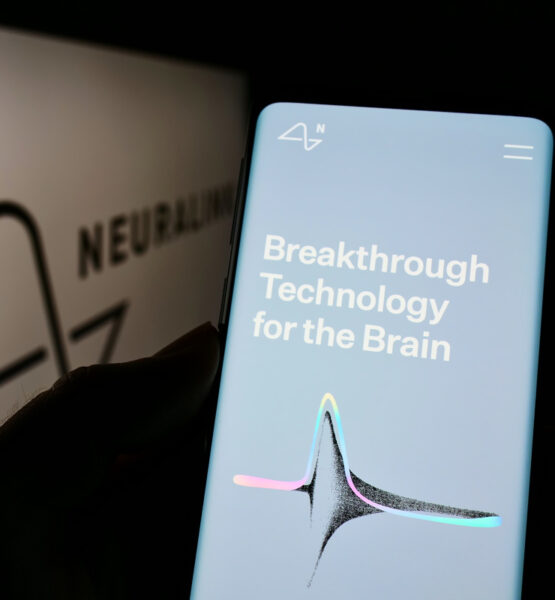 Ein Smartphone wird in der Hand gehalten und zeigt eine Anzeige mit dem Text "Breakthrough Technology for the Brain". Im Hintergrund ist das Logo von Neuralink, einem Unternehmen, das sich auf die Entwicklung von Neurotechnologie spezialisiert hat, teilweise sichtbar. Das Logo erscheint unscharf und hinter dem Smartphone, was darauf hindeutet, dass der Fokus auf dem Bildschirm des Smartphones liegt. Der Bildschirm ist hellblau mit weißem Text und einem grafischen Element in der Mitte, das eine stilisierte Darstellung einer Neuronen- oder Synapsenstruktur sein könnte, mit Farbakzenten in Rosa und Grün an den Enden.