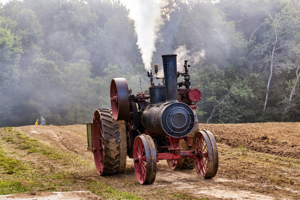 Ein alter Dampftraktor mit großen Metallrädern steht auf einem frisch gepflügten Feld. Aus dem Schornstein des Traktors steigt Rauch auf. Im Hintergrund sieht man Bäume und es ist ein leichter Nebel zu erkennen.
