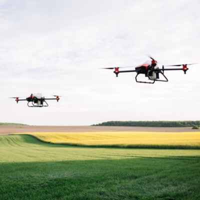 Bild 1: Zwei Drohnen fliegen nebeneinander über eine landwirtschaftliche Fläche mit unterschiedlichen Anbauflächen. Der Himmel ist klar und es gibt eine sichtbare Trennung zwischen den grünen und gelbbraunen Feldern im Hintergrund.