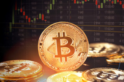 Das Bild zeigt physische Bitcoin-Münzen im Vordergrund mit einer unscharfen Anzeige eines Börsenkurs-Diagramms im Hintergrund. Die Münzen sind repräsentativ für die Kryptowährung Bitcoin und scheinen auf die Verbindung zwischen digitalen Währungen und Finanzmärkten hinzuweisen. Das Diagramm im Hintergrund könnte auf die Volatilität und Handelsaktivität von Bitcoin oder anderen Kryptowährungen auf den Märkten hindeuten.