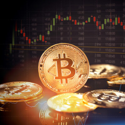 Das Bild zeigt physische Bitcoin-Münzen im Vordergrund mit einer unscharfen Anzeige eines Börsenkurs-Diagramms im Hintergrund. Die Münzen sind repräsentativ für die Kryptowährung Bitcoin und scheinen auf die Verbindung zwischen digitalen Währungen und Finanzmärkten hinzuweisen. Das Diagramm im Hintergrund könnte auf die Volatilität und Handelsaktivität von Bitcoin oder anderen Kryptowährungen auf den Märkten hindeuten.
