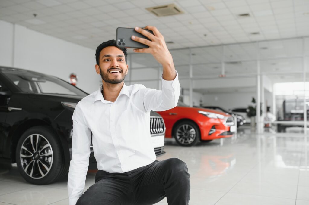 Ein junger Mann in einem weißen Hemd macht ein Selfie mit seinem Smartphone in einer hellen Autoausstellungshalle. Er sitzt und lächelt, während im Hintergrund verschiedene Autos zu sehen sind.