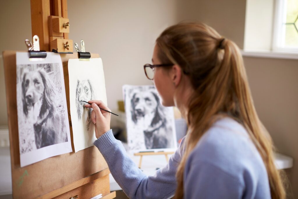 Eine junge Frau mit Brille und zurückgebundenen blonden Haaren sitzt an einem Holzstaffelei und zeichnet das Porträt eines Hundes. Neben ihr auf der Staffelei ist ein Schwarz-Weiß-Foto des Hundes zu sehen, welches als Vorlage für die Zeichnung dient.