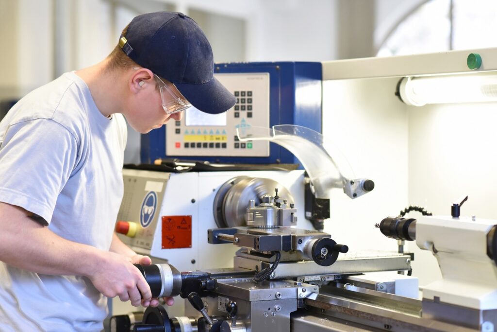 Ein junger Mann mit einer Kappe und Schutzbrille arbeitet konzentriert an einer metallbearbeitenden Drehmaschine. Er trägt ein weißes T-Shirt und bedient die Maschine in einer industriellen Werkstattumgebung.