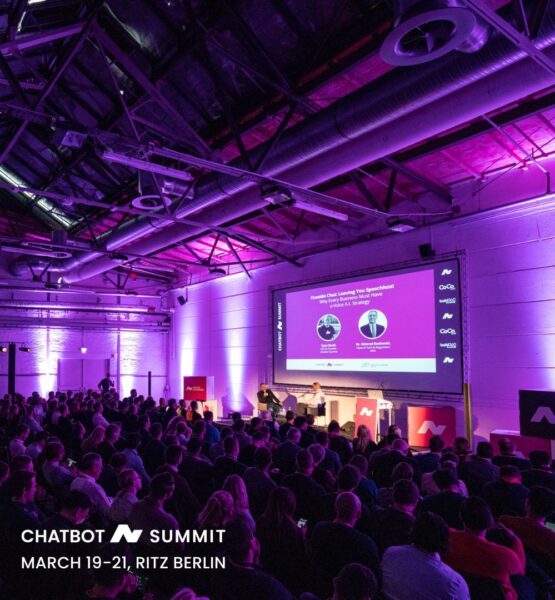 Blick auf eine von violettem Licht beleuchtete Bühne des Chatbot Summit, auf der ein Fireside-Chat vor großen Publikum stattfindet.