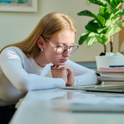 Eine junge Frau mit langen blonden Haaren und Brille sitzt nachdenklich vor ihrem Laptop, gestützt auf einen weißen Schreibtisch. Ein Stapel von Papieren und eine grüne Zimmerpflanze sind im Hintergrund sichtbar.