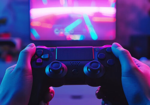 Gaming-Controller in der Hand und im Hintergrund ein Bildschirm auf dem ein verschwommenes Videospiel angezeigt wird. Es soll Mikrotransaktionen im Gaming darstellen.