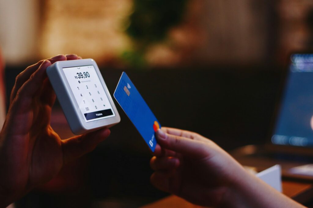 Nahaufnahme einer Hand, die ein tragbares Kartenlesegerät mit einem Touchscreen hält, auf dem ein Preis angezeigt wird. Eine andere Hand hält eine blaue Kreditkarte in Vorbereitung darauf, die Zahlung abzuschließen. Der Fokus liegt auf dem Austausch und der Technologie, die im modernen Zahlungsverkehr verwendet wird.