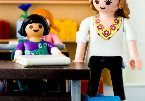 Unterrichtsszene mit einer Lehrerin, die bei einer Schülerin mit Arbeitslaptop steht mit Playmobil nachgestellt.