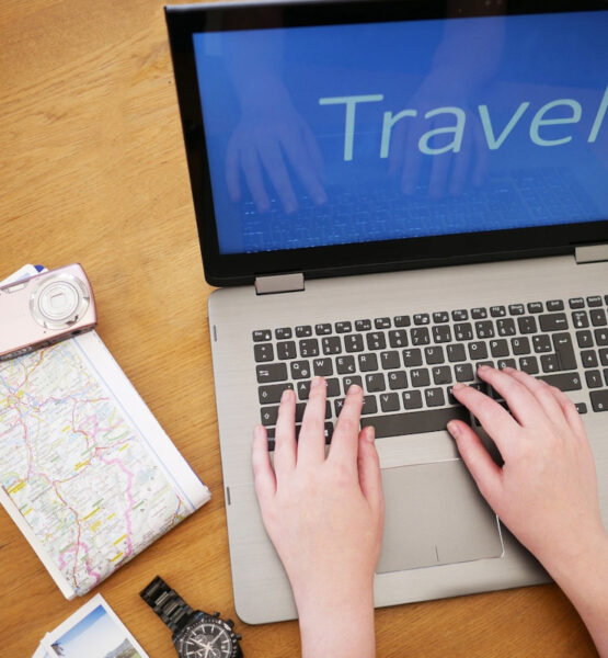 Ein Arbeitsplatz mit einem geöffneten Laptop, der das Wort „Travel“ auf dem Bildschirm zeigt. Auf dem Tisch liegen neben dem Laptop eine Landkarte, eine Kamera, ein paar Fotos und eine Armbanduhr. Die Person tippt auf der Tastatur, was darauf hinweist, dass sie eine Reise plant oder Informationen über Reiseziele recherchiert.