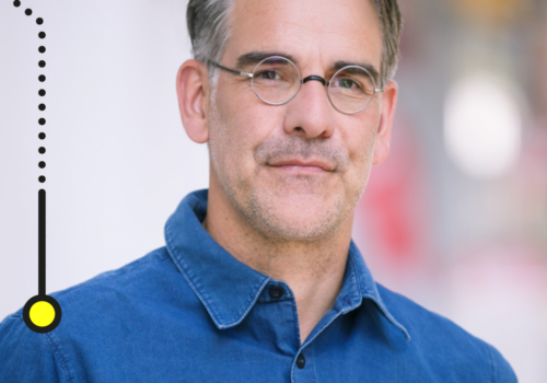 Das Bild zeigt einen Mann mittleren Alters mit graumelierten Haaren und einem gepflegten Bart. Er trägt eine runde Brille und ein blaues Hemd. Der Hintergrund ist verschwommen, wodurch der Fokus auf dem Mann liegt. In der oberen linken Ecke befindet sich ein Logo mit der Aufschrift "brand eins Netzwerk".