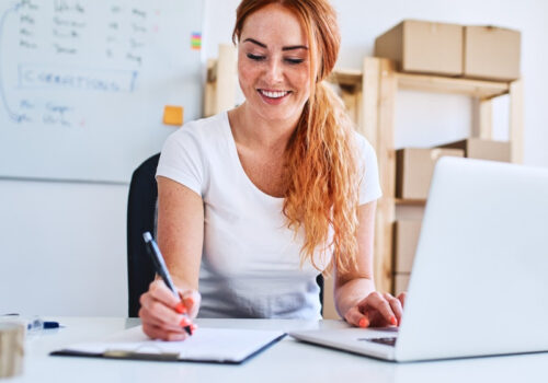 Eine junge Geschäftsinhaberin sitzt im Büro und schreibt Verkaufs- und Lieferdaten auf Papier, während sie einen Laptop benutzt.