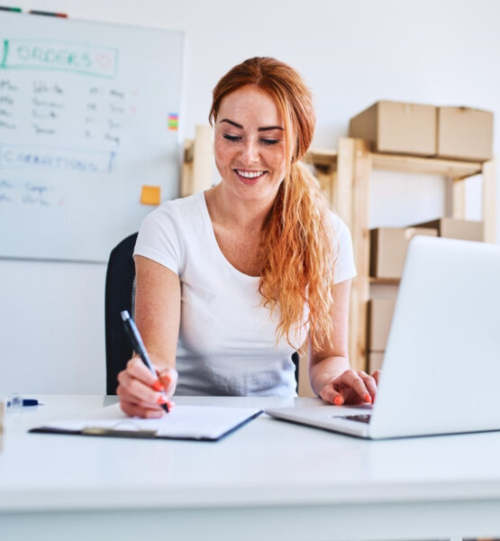 Eine junge Geschäftsinhaberin sitzt im Büro und schreibt Verkaufs- und Lieferdaten auf Papier, während sie einen Laptop benutzt.