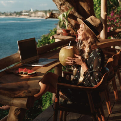 Eine junge Frau sitzt auf einer Holzterrasse mit Blick auf das Meer. Sie trägt einen Strohhut und ein florales Kleid und hält eine Kokosnuss mit Strohhalm in der Hand. Vor ihr auf dem Tisch steht ein Laptop und eine Platte mit Wassermelonenstücken. Sie scheint von einem Urlaubsort aus entspannt zu arbeiten.
