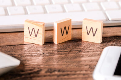 Nahaufnahme eines Schreibtischs mit einer weißen Computertastatur im Hintergrund. Im Vordergrund befinden sich drei kleine mit Buchstaben beschriftete Holzplättchen, die den Begriff "WWW" ergeben. Links ist eine weiße Computermaus zu sehen, und rechts ein Smartphone mit schwarzem Bildschirm. Der Schreibtisch ist aus Holz und die Szene wirkt ordentlich und aufgeräumt.