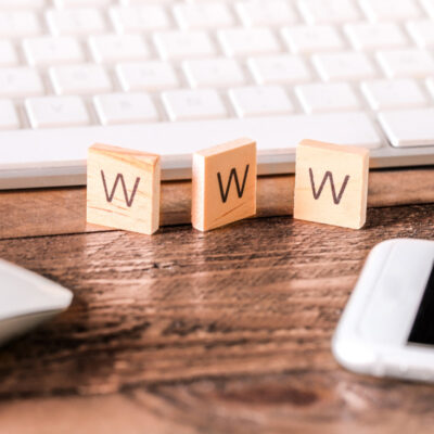 Nahaufnahme eines Schreibtischs mit einer weißen Computertastatur im Hintergrund. Im Vordergrund befinden sich drei kleine mit Buchstaben beschriftete Holzplättchen, die den Begriff "WWW" ergeben. Links ist eine weiße Computermaus zu sehen, und rechts ein Smartphone mit schwarzem Bildschirm. Der Schreibtisch ist aus Holz und die Szene wirkt ordentlich und aufgeräumt.