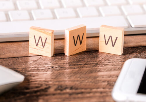 Nahaufnahme eines Schreibtischs mit einer weißen Computertastatur im Hintergrund. Im Vordergrund befinden sich drei kleine mit Buchstaben beschriftete Holzplättchen, die den Begriff "WWW" ergeben. Links ist eine weiße Computermaus zu sehen, und rechts ein Smartphone mit schwarzem Bildschirm. Der Schreibtisch ist aus Holz und die Szene wirkt ordentlich und aufgeräumt.