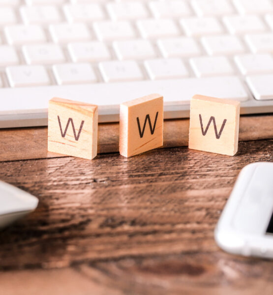 Nahaufnahme eines Schreibtischs mit einer weißen Computertastatur im Hintergrund. Im Vordergrund befinden sich drei kleine mit Buchstaben beschriftete Holzplättchen, die den Begriff "WWW" ergeben. Links ist eine weiße Computermaus zu sehen, und rechts ein Smartphone mit schwarzem Bildschirm. Der Schreibtisch ist aus Holz und die Szene wirkt ordentlich und aufgeräumt.