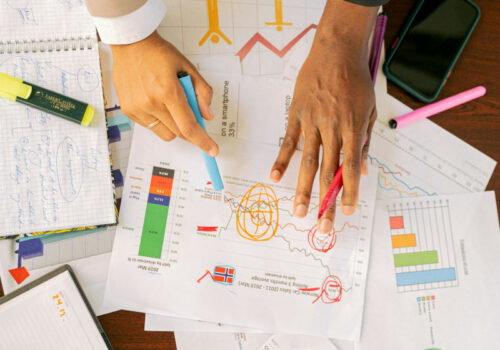 Eine Nahaufnahme von Händen, die auf verschiedenen Geschäftsdokumenten und Diagrammen arbeiten. Die Hände halten bunte Stifte und markieren oder notieren Informationen auf den Dokumenten. Um die Dokumente herum liegen Notizbücher, ein Smartphone und verschiedene Büroartikel. Die Szene vermittelt eine konzentrierte Arbeitsatmosphäre, in der Daten analysiert und Strategien entwickelt werden.