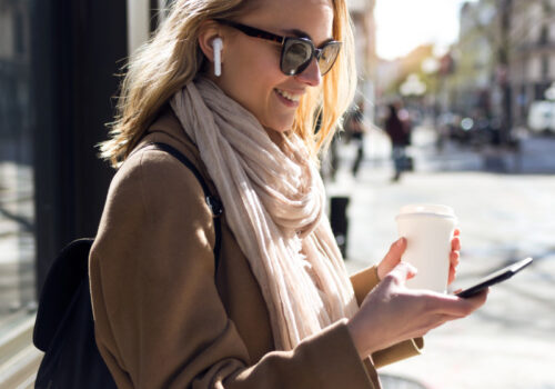 Eine junge Frau steht draußen in einer städtischen Umgebung und lächelt, während sie auf ihr Smartphone schaut. Sie trägt Sonnenbrillen, kabellose Ohrhörer, einen beigen Mantel und einen hellen Schal. In der linken Hand hält sie einen Kaffeebecher. Im Hintergrund sind verschwommen Gebäude und Fußgänger zu sehen, die eine belebte Straßenszene andeuten. Die Frau wirkt zufrieden und beschäftigt sich vermutlich mit einer Nachricht oder einem Anruf auf ihrem Smartphone.