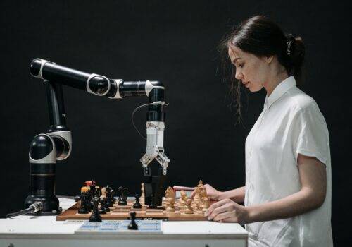 Eine Frau in einem weißen Hemd spielt Schach gegen einen Roboterarm. Der Roboter greift mit seiner mechanischen Hand nach einer Schachfigur auf einem Schachbrett. Die Szene symbolisiert den Wettstreit zwischen menschlicher Intuition und maschineller Präzision. Der Hintergrund ist dunkel, was die beiden Spieler hervorhebt.