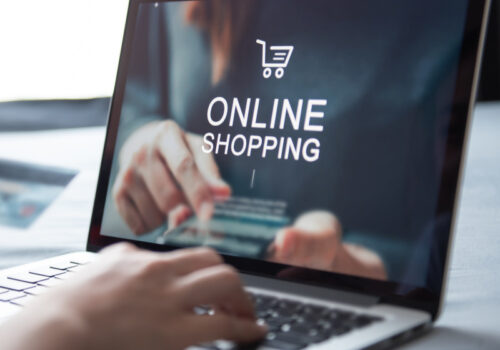 Eine Person nutzt einen Laptop für Online-Shopping, hält eine Kreditkarte in der Hand und tippt auf der Tastatur. Auf dem Bildschirm ist der Schriftzug 'Online Shopping' mit einem Einkaufswagensymbol zu sehen. Im Hintergrund befindet sich eine unscharfe, dunkle Umgebung.