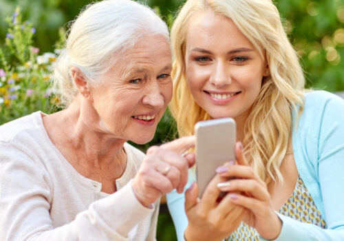 "Eine ältere Frau und eine jüngere Frau sitzen gemeinsam draußen an einem Tisch. Sie lächeln, während sie gemeinsam auf ein Smartphone schauen. Auf dem Tisch stehen eine Tasse, ein Teller mit Kuchen und ein Blumenarrangement im Hintergrund."