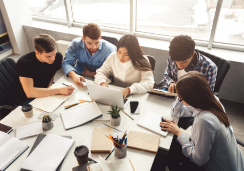 Eine Gruppe von Studenten arbeitet gemeinsam an einem Tisch mit Laptops. Sie sind in eine konzentrierte Teamarbeit vertieft und umgeben von Schreibmaterialien wie Stifte, Notizbücher und Ordner. Die Szene vermittelt ein produktives Lernumfeld mit moderner Technologie.